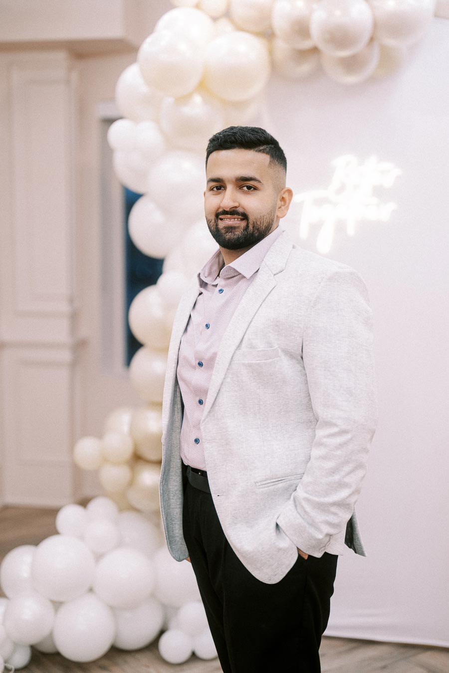A man in a light gray blazer and pink shirt smiles at a decorated event venue with white balloons in the background.