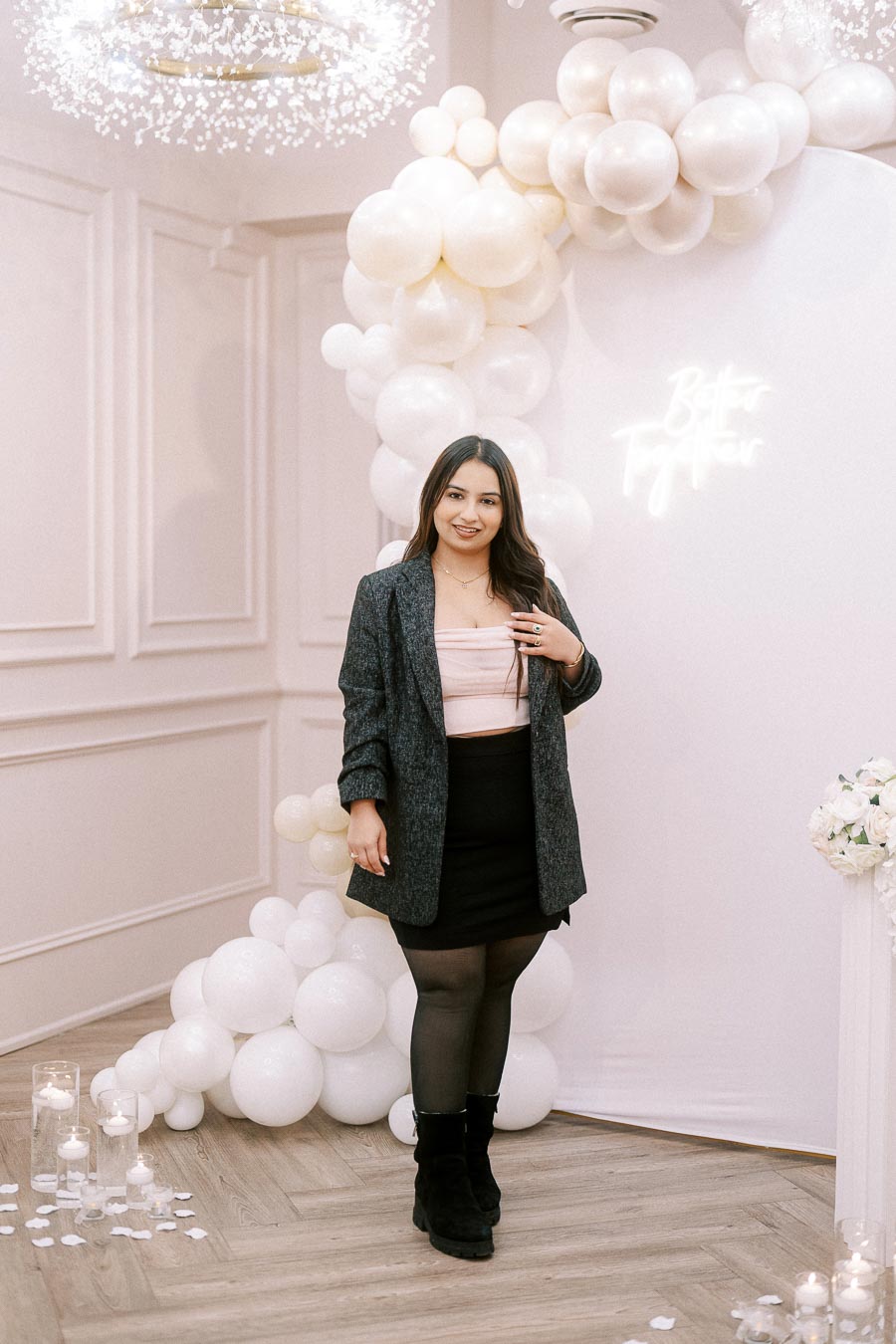 A woman in a stylish outfit stands in a decorated room with white balloon arch and a glowing Better Together sign, exuding elegance at a chic event.