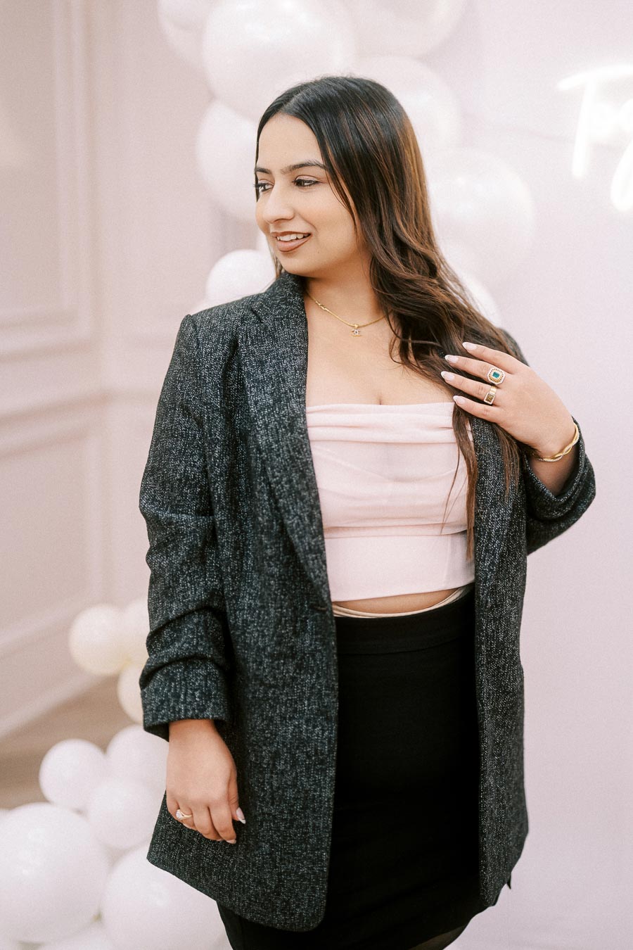 A stylish woman in a gray blazer and pink top poses confidently in front of a soft, elegant backdrop with white balloons.