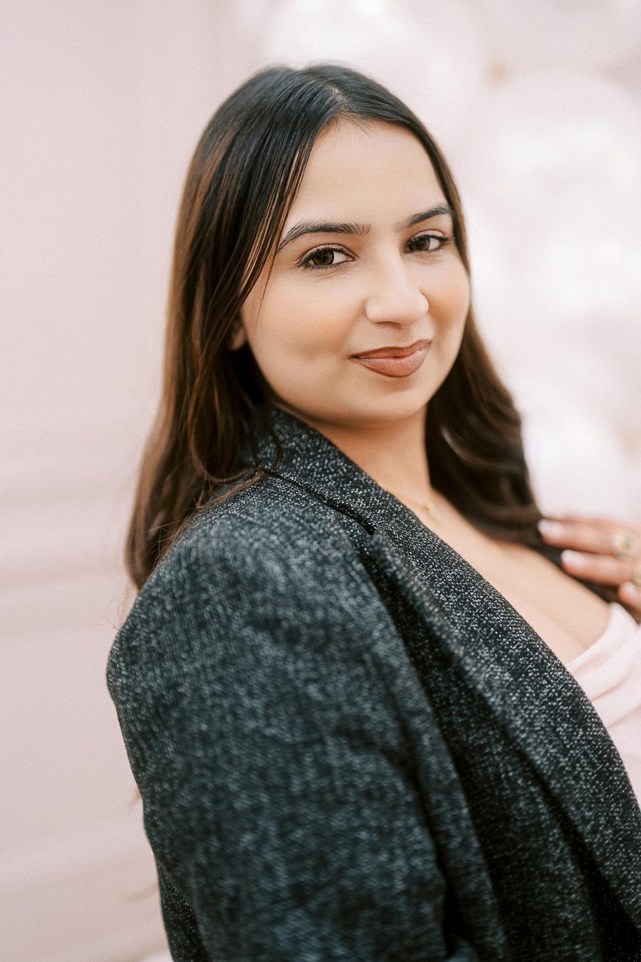 Young woman smiling confidently, wearing a textured grey blazer over a light pink top, with a soft pink and white background.