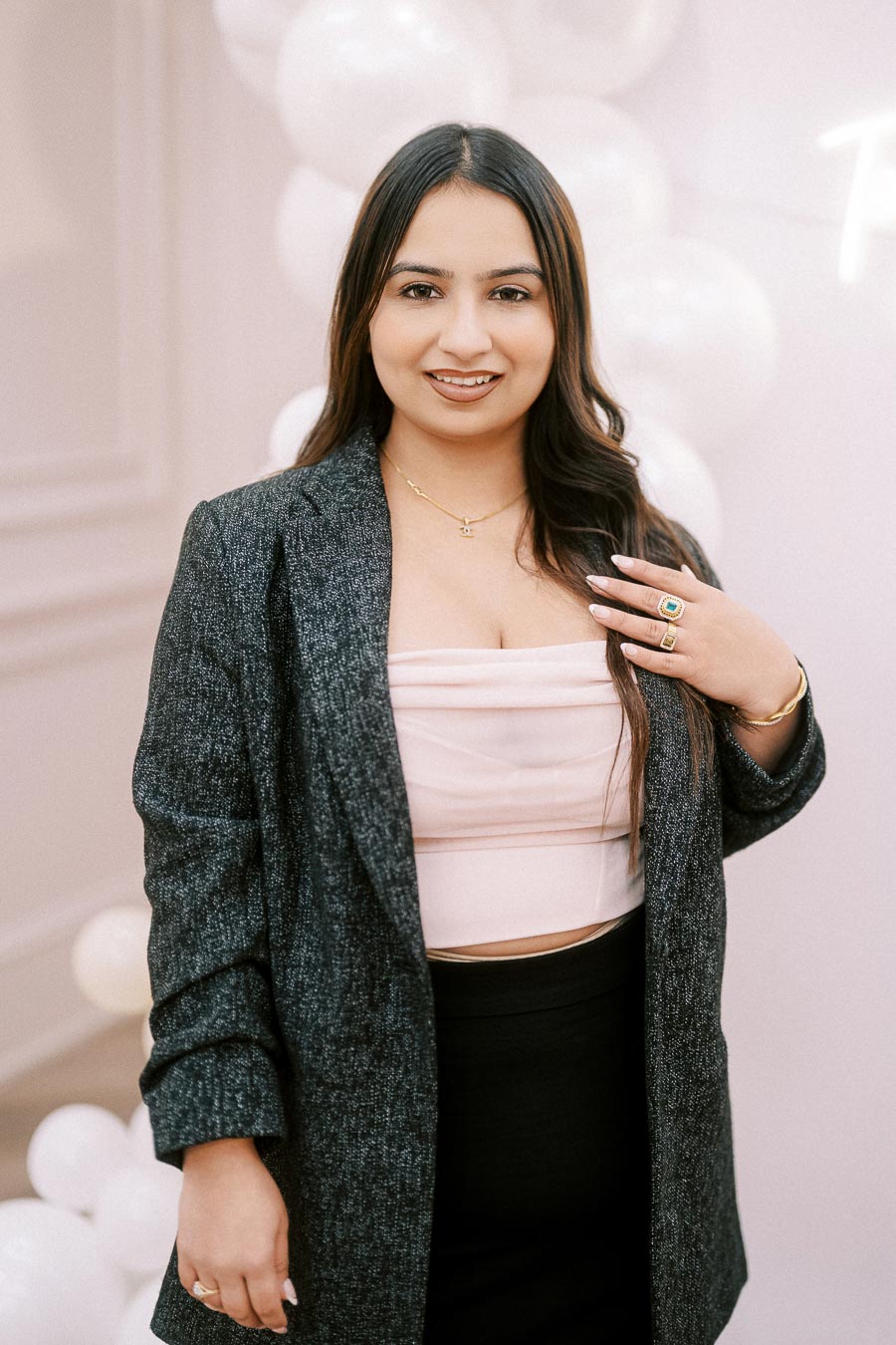 A confident woman smiling at the camera, wearing a gray blazer and pink top, with long dark hair and accessorized with rings and a necklace, standing in front of a white balloon backdrop.