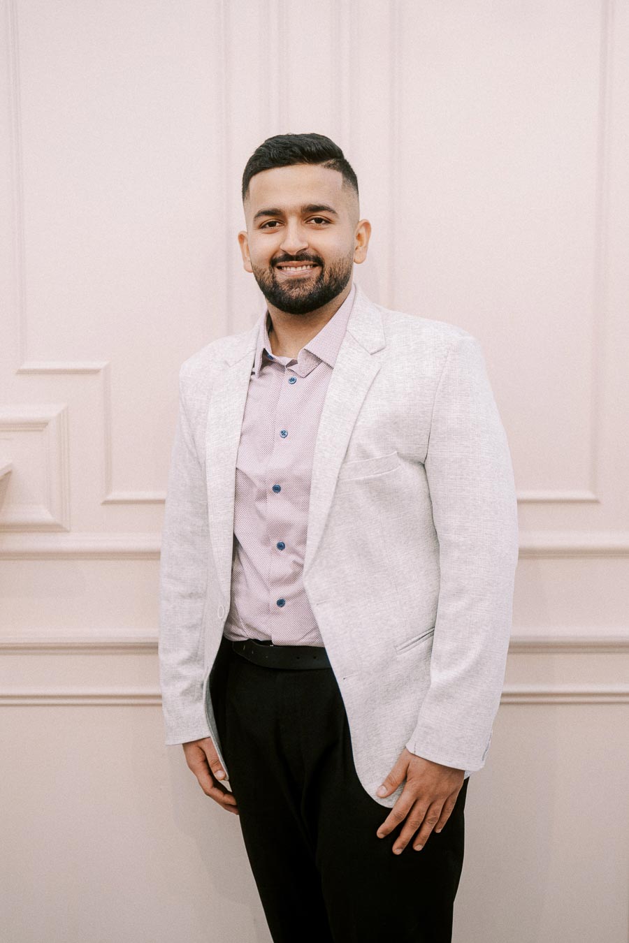 A man in a light grey blazer and pink shirt smiling against a light-colored, textured background.