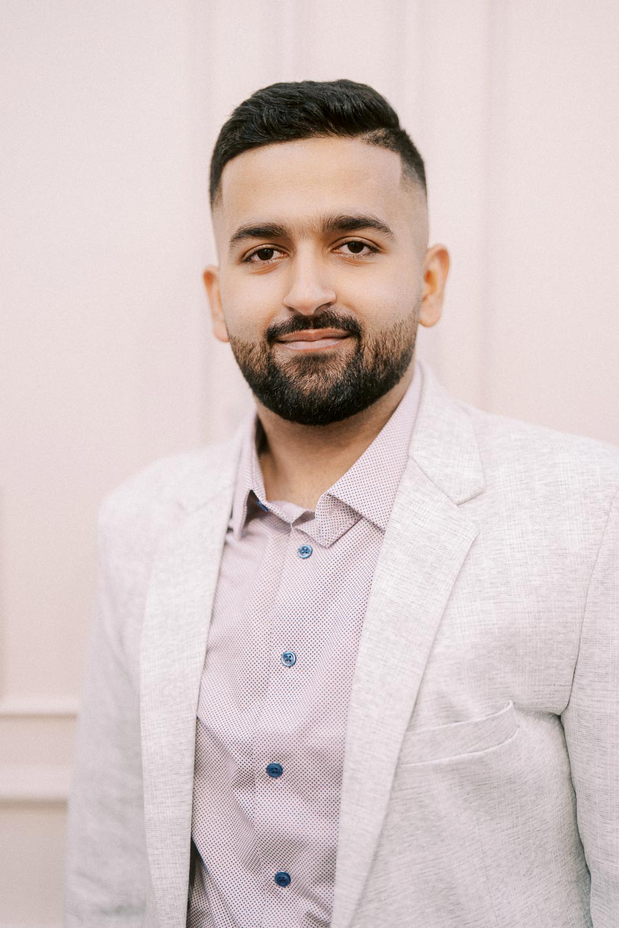 A well-dressed man with a neat beard, wearing a light gray blazer over a patterned shirt, poses confidently against a neutral background.
