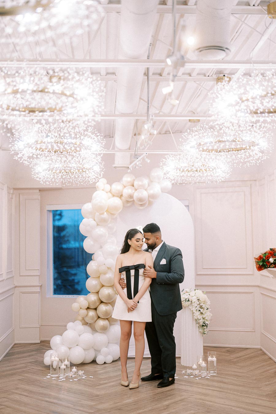 A stylish couple posing in an elegantly decorated room with white and gold balloons, under sparkling chandeliers, dressed in formal attire, ideal for a sophisticated engagement or wedding party setting.