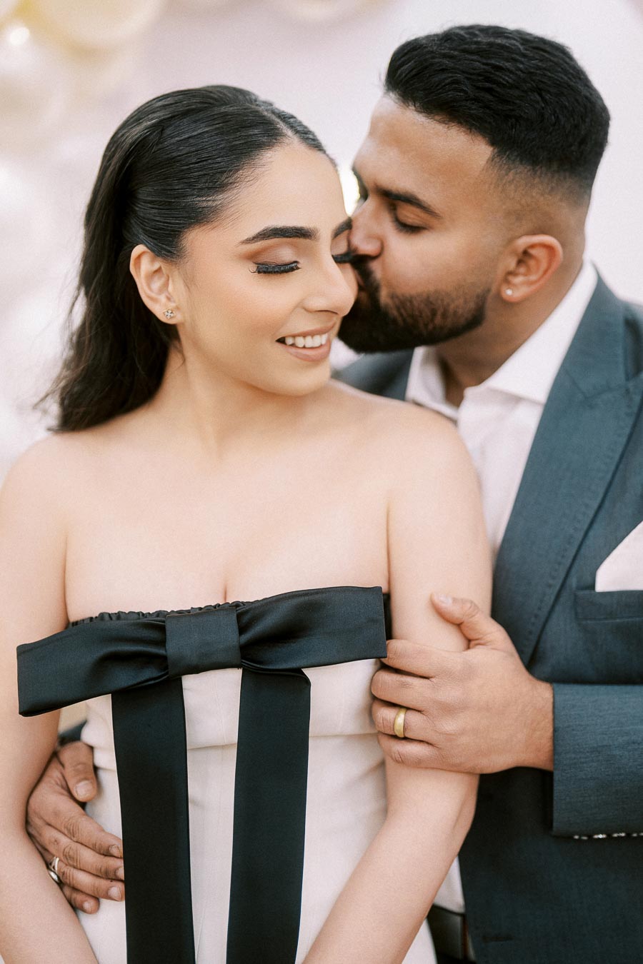 A loving couple embracing; the man kisses the woman on the cheek while gently holding her. The woman smiles, wearing a strapless dress with a large black bow, creating a romantic and elegant atmosphere.