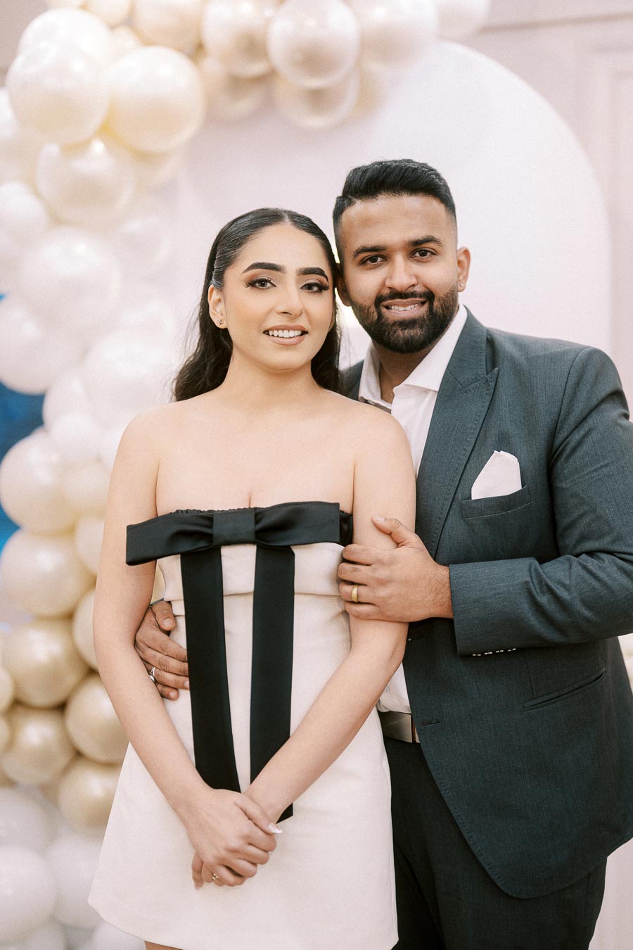Alt text: Smiling couple posing in elegant attire at a formal event, with a decorative balloon arch in the background.