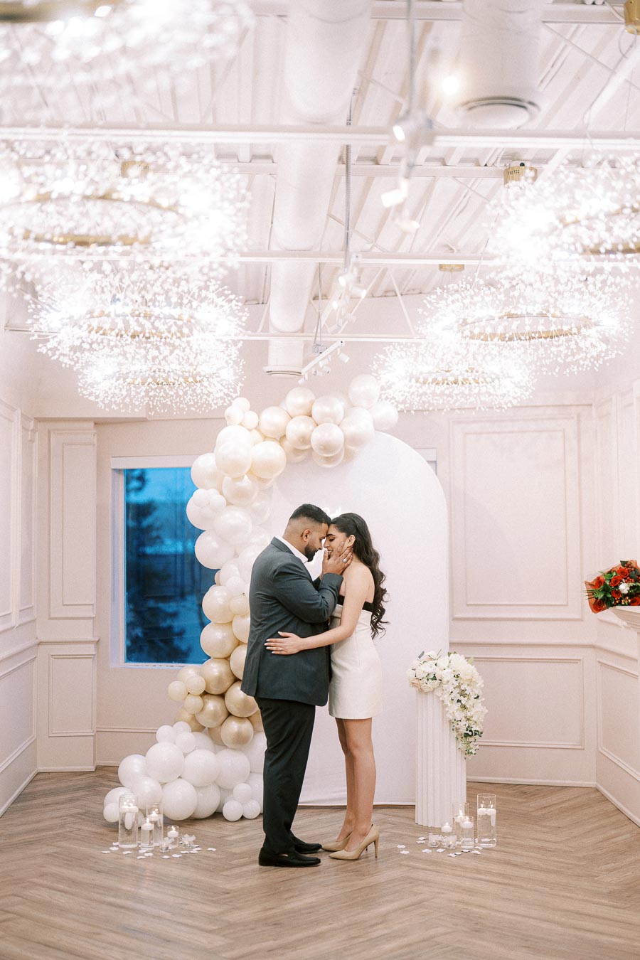 A couple embracing in an elegant room with a romantic balloon arch, floral arrangements, and ambient lighting, perfect for an intimate wedding celebration.