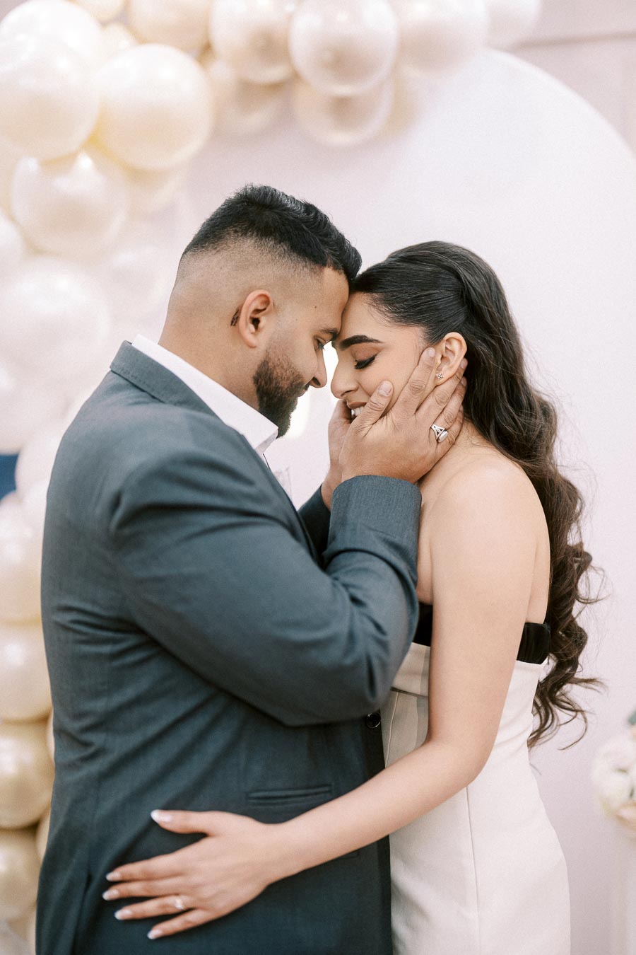 A couple embracing with foreheads touching, surrounded by elegant white balloons, captured in a tender and romantic moment.