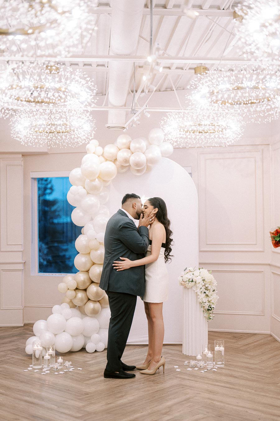 A couple shares a romantic moment surrounded by an elegant wedding decor, featuring a stunning backdrop of white and gold balloons and delicate floral arrangements, in a beautifully lit venue.