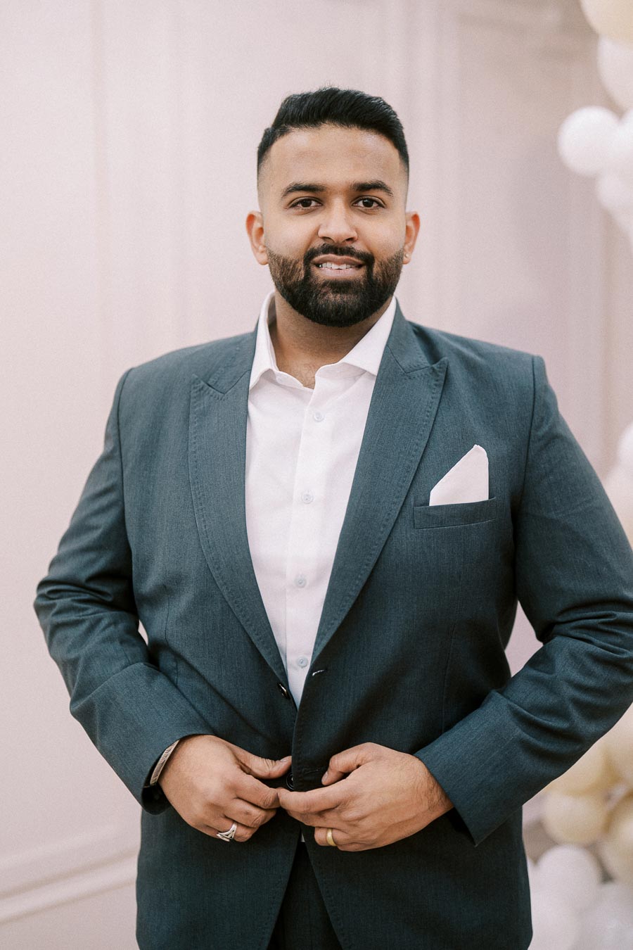 A man in a grey suit with a white shirt stands against a light background, confidently looking at the camera. The suit features a pocket square and he is adjusting the buttons.