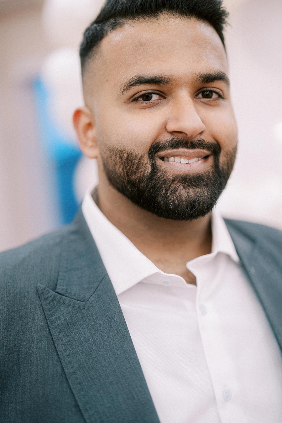 Smiling man wearing a grey suit and white shirt at an event with blurred background.