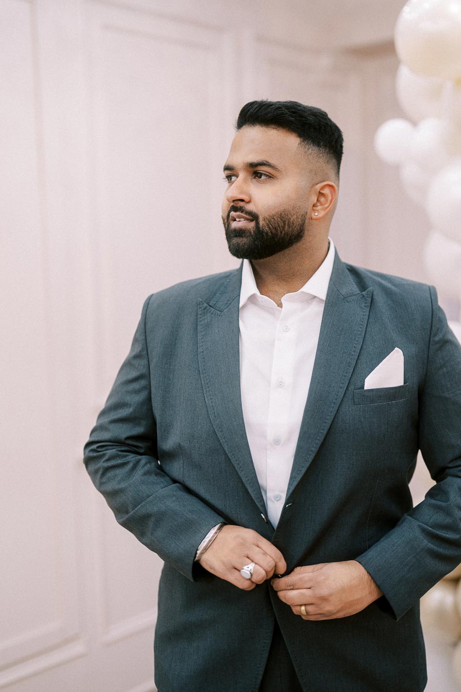 A man with a neatly groomed beard wearing a stylish gray suit and white shirt, adjusting his jacket in a sophisticated setting with soft lighting and decorative balloons in the background.
