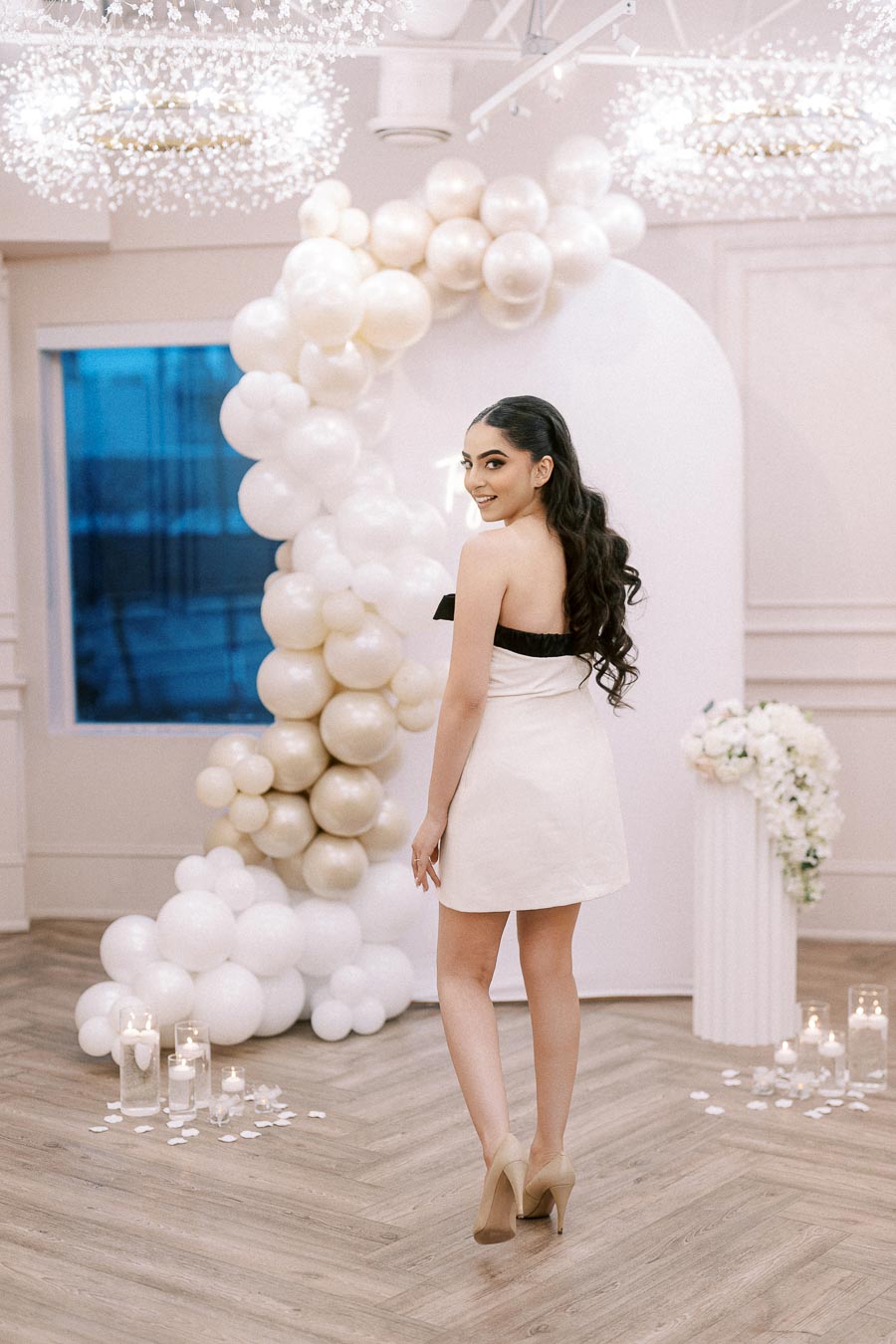 Elegant woman in a strapless white dress posing in a decorated room with pearl and white balloon arch, surrounded by candles and floral arrangements.
