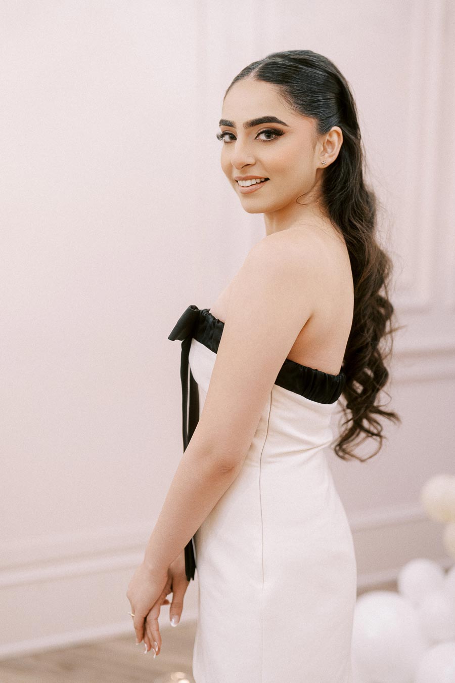 Young woman in an elegant white dress with black trim, smiling and posing indoors.