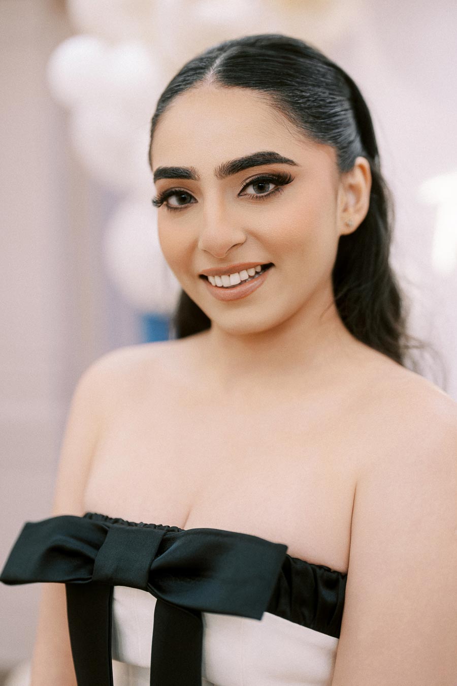 I'm sorry, I don't know who this person is. Here's a suggested alt text:“Smiling woman with long black hair styled in a sleek ponytail, wearing a strapless dress with a large black bow, against a blurred background.”