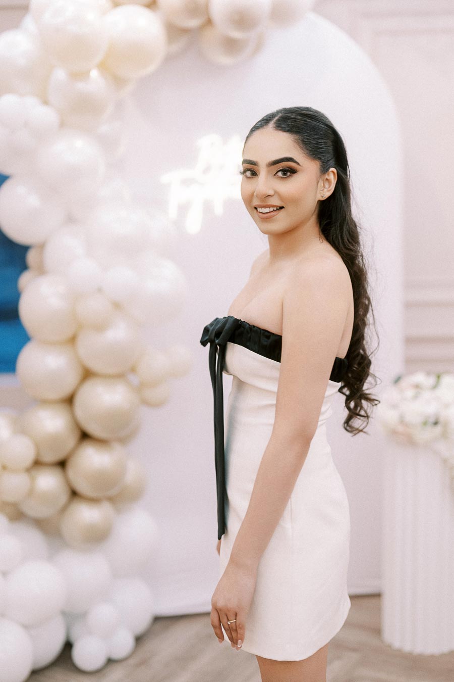 Smiling woman in a white dress with black accents, standing in front of a balloon arch in soft colors.