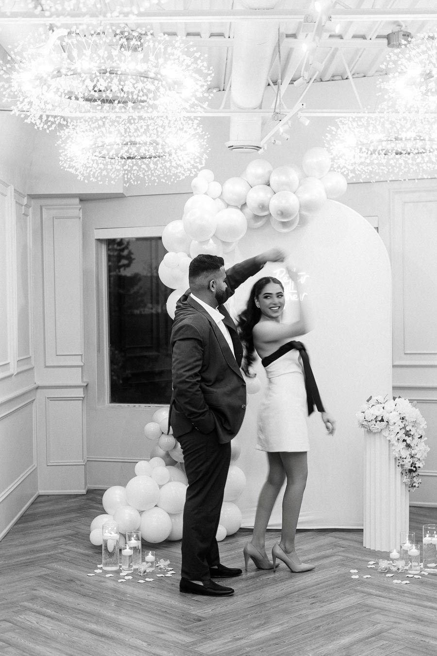 A joyful couple dancing in a beautifully decorated room with elegant lighting and white balloon arch decor. The woman is wearing a chic white dress and high heels, while the man is dressed in a formal suit. The atmosphere is festive, with scattered rose petals and candles enhancing the romantic setting.