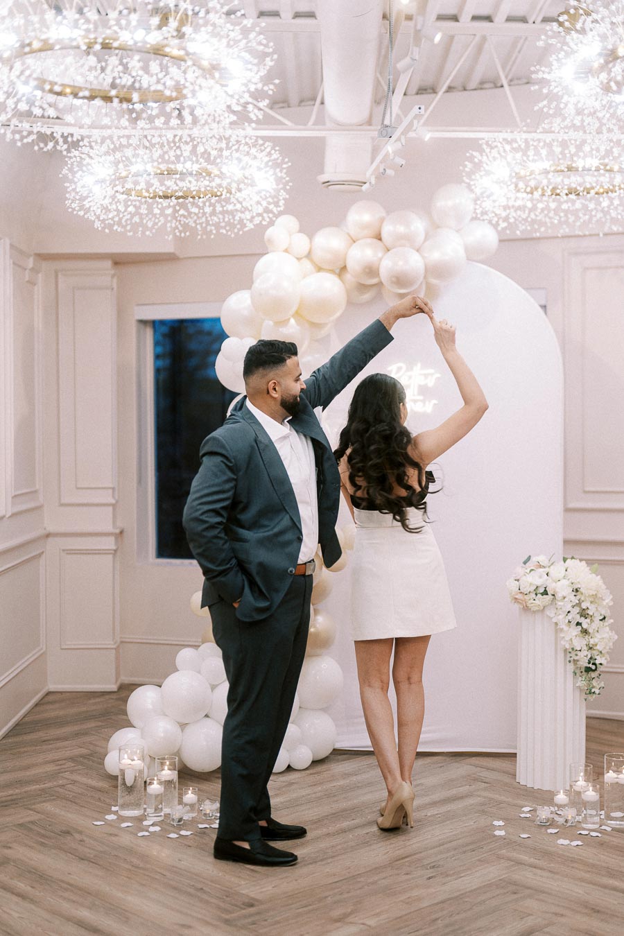 A couple dances under elegant lighting with a balloon arch and candles, creating a romantic atmosphere in a stylishly decorated room.