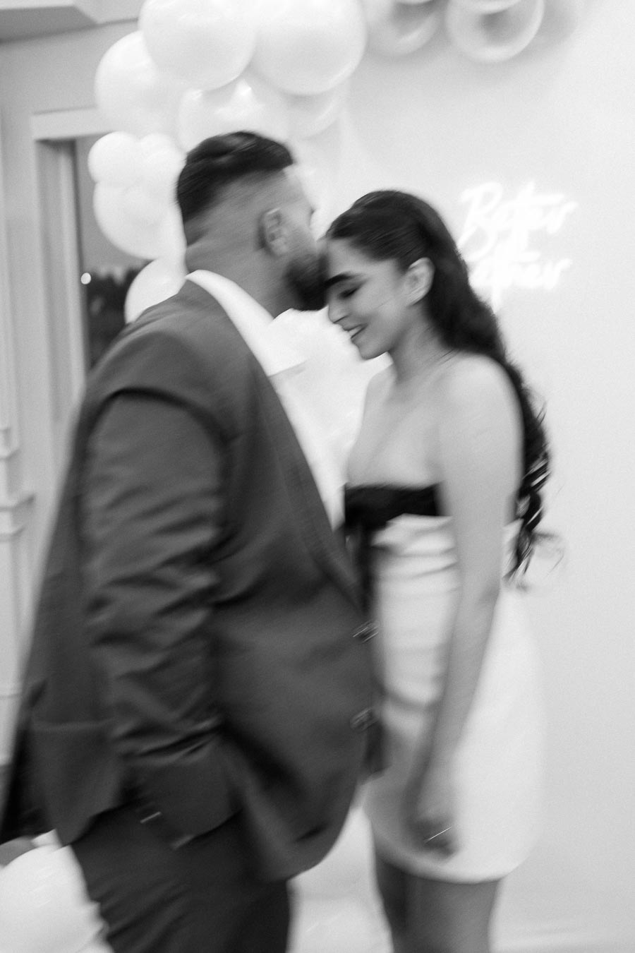 A black and white photo of a couple in formal attire sharing an intimate moment, with a background decorated with white balloons.