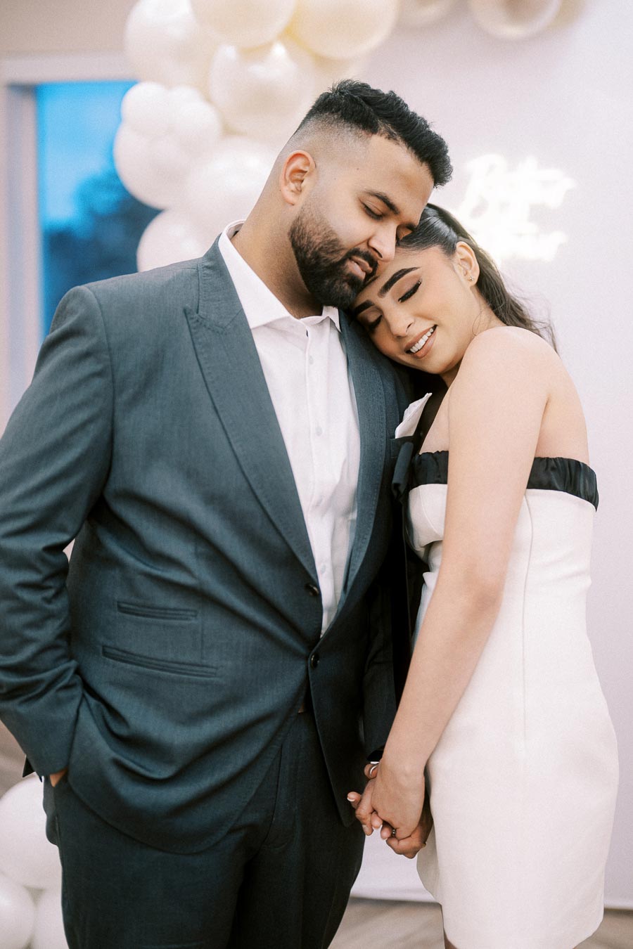 A couple smiling and embracing each other at an elegant celebration, surrounded by white balloons and dressed in formal attire, creating a romantic atmosphere.