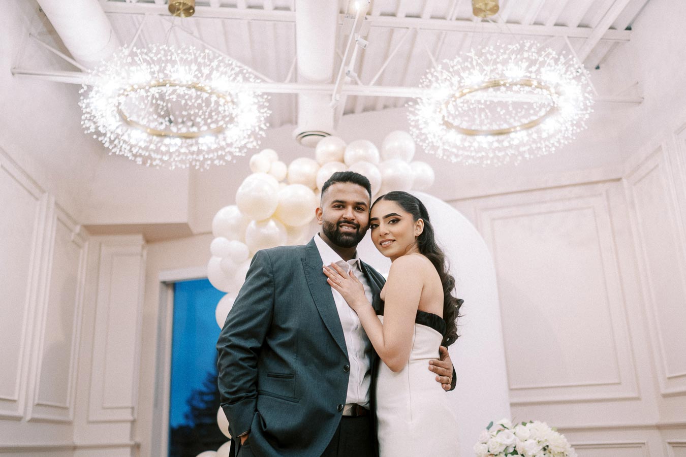 A couple in formal attire poses indoors under elegant chandeliers, with a backdrop of white balloons, exuding a romantic and celebratory atmosphere.
