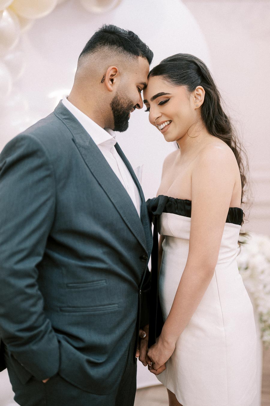 Happy couple embracing and smiling at each other, dressed in formal attire with a romantic balloon backdrop, ideal for wedding or engagement themes.
