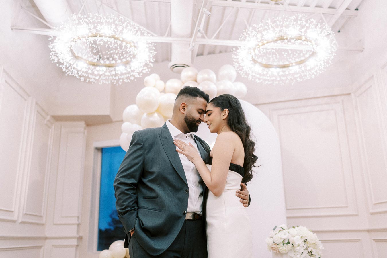 A romantic couple embraces in an elegant setting with soft lighting and white floral decorations, under modern circular chandeliers.