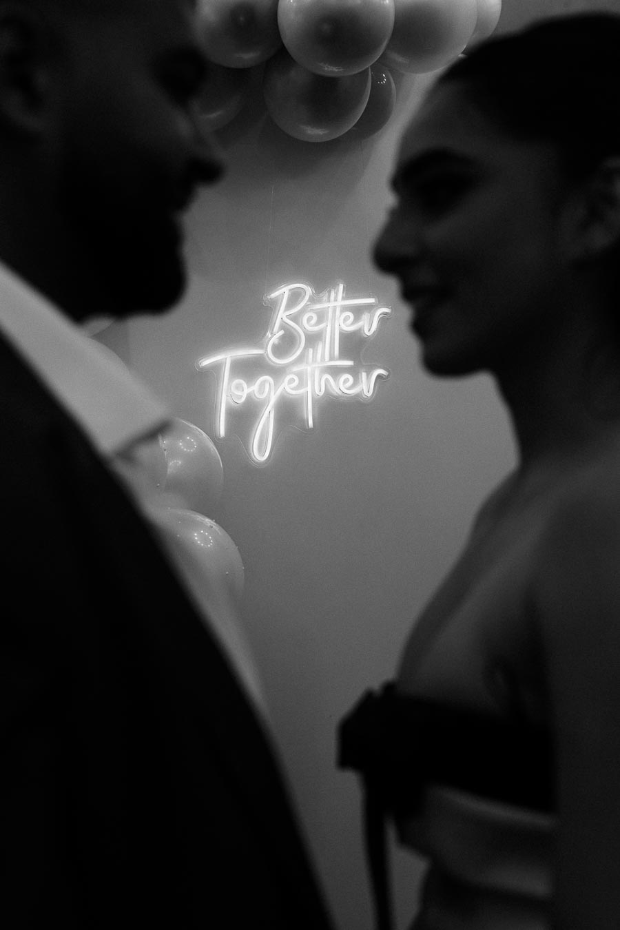 Couple in silhouette facing each other with Better Together neon sign and balloons in background, black and white.