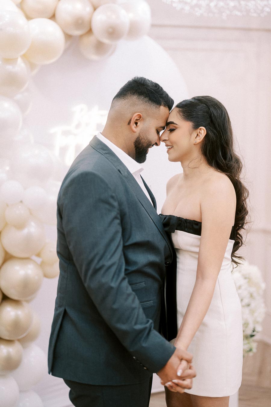 Happy couple sharing a tender moment at their engagement celebration, stylishly dressed with elegant balloon decorations in the background.