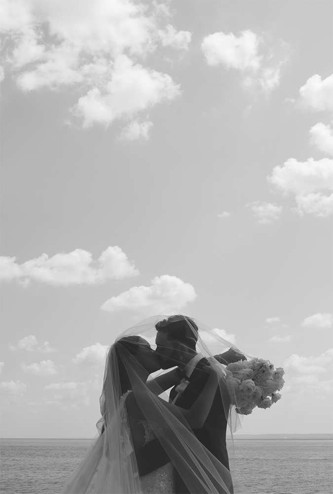 Black and white image of a romantic wedding kiss by the sea, with a bride and groom embracing under a veil against a cloudy sky backdrop.