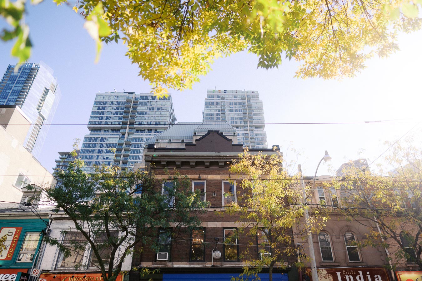 Street view of a historic brick building in front of modern glass skyscrapers, with autumn tree foliage and a clear blue sky in an urban setting.