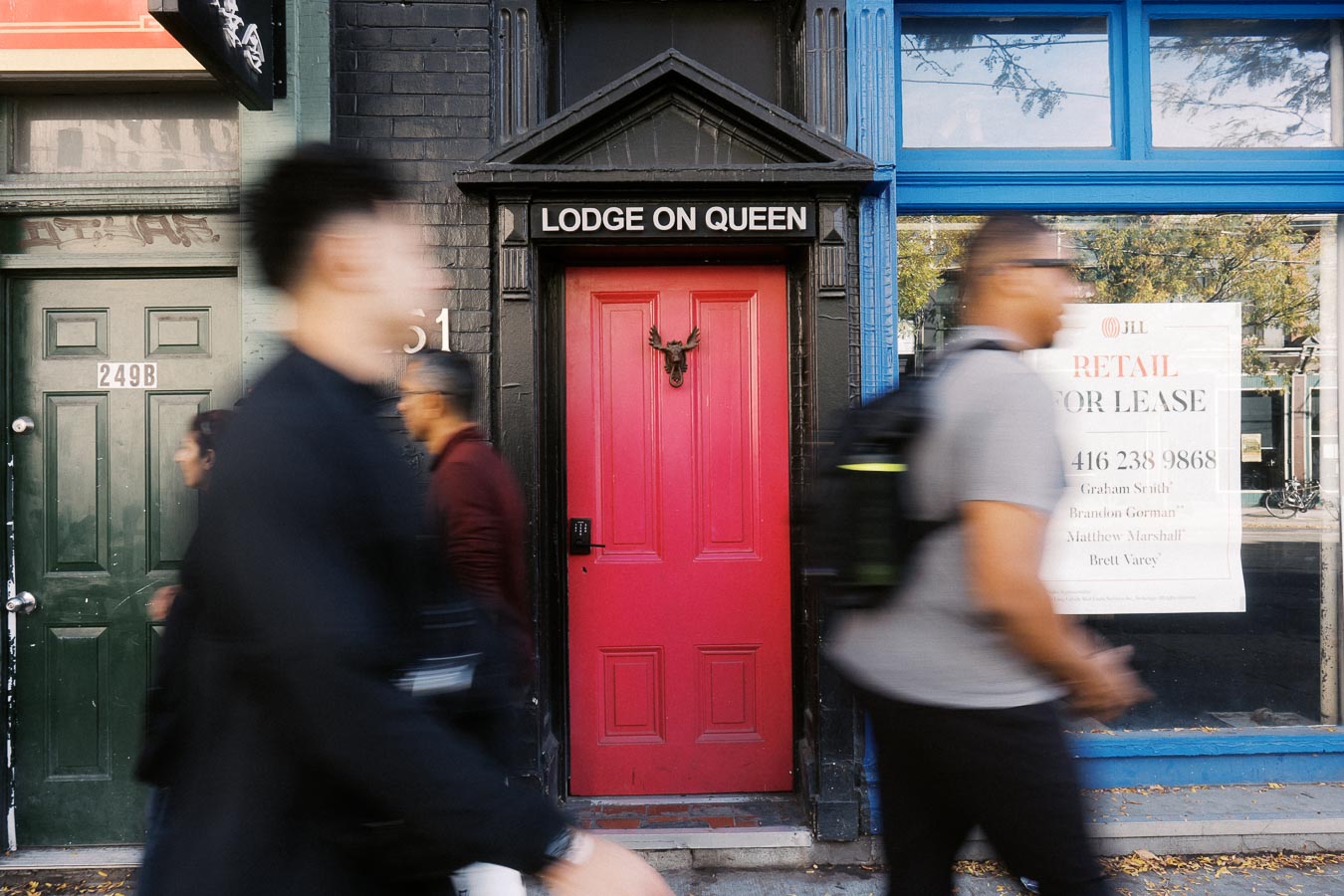 Alt text: Street view of a vibrant urban scene featuring a bright red door labeled 'Lodge on Queen' with blurred pedestrians walking by, next to a retail space for lease.