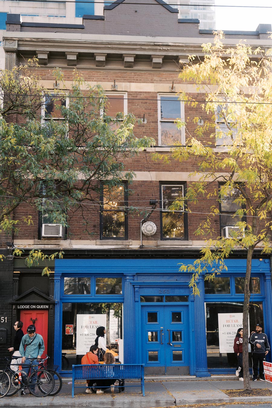 Street view of a charming urban building with a bright blue facade, featuring a Retail for Lease sign on the window. A group of people sit on a blue bench outside, with bicycles parked nearby. Autumn trees add a touch of nature to the bustling city scene.