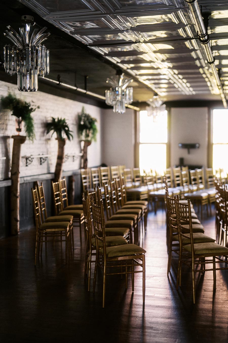 Elegant event space with rows of golden chairs and lush greenery, illuminated by natural light and sparkling chandeliers.