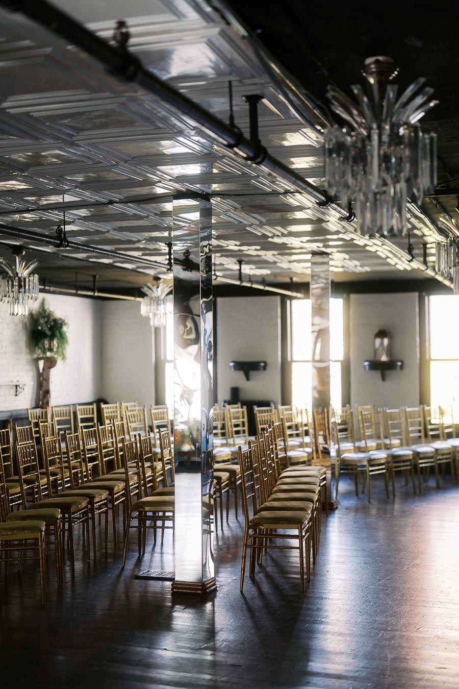 Elegant event space with rows of gold chairs, ornate chandeliers, mirrored columns, and natural light filtering through windows.