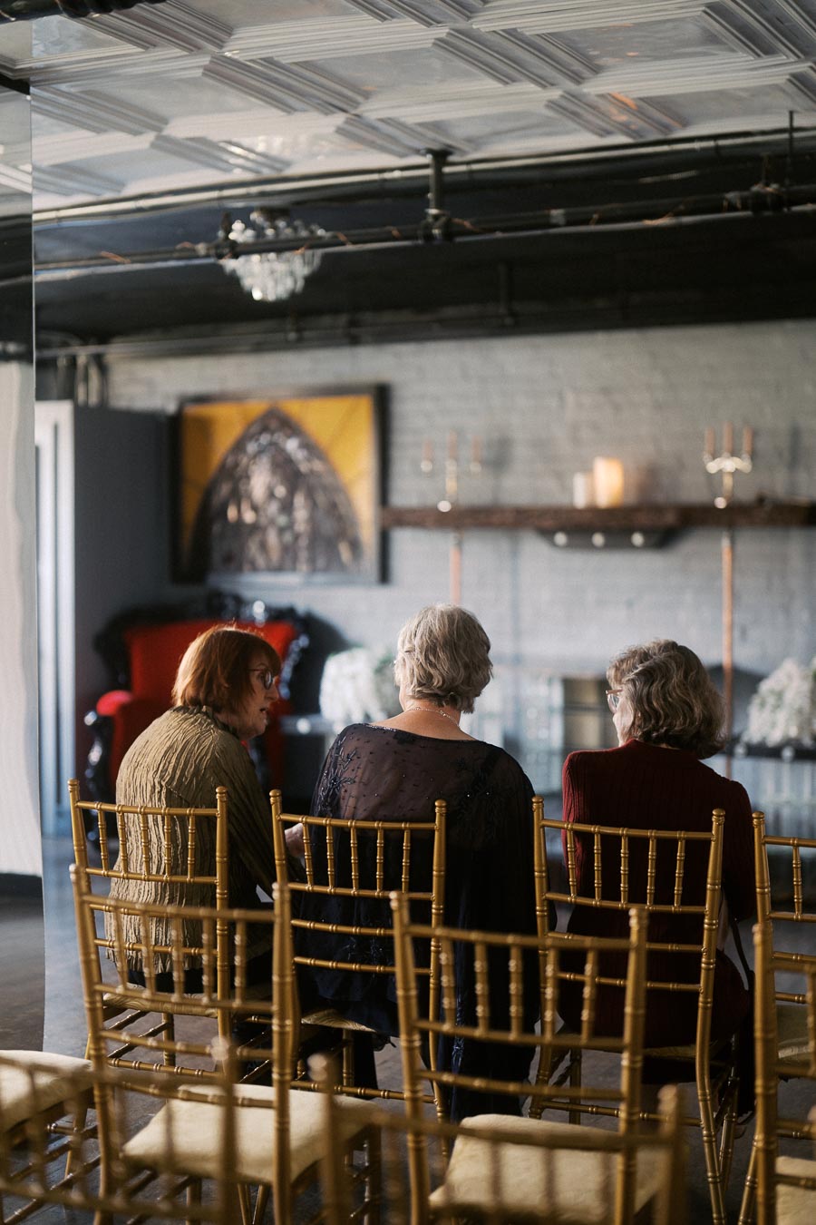A group of three women sitting on gold chairs in an elegant room, engaged in conversation. The background features a red chair, decorative wall art, and candles, creating a sophisticated and intimate atmosphere.