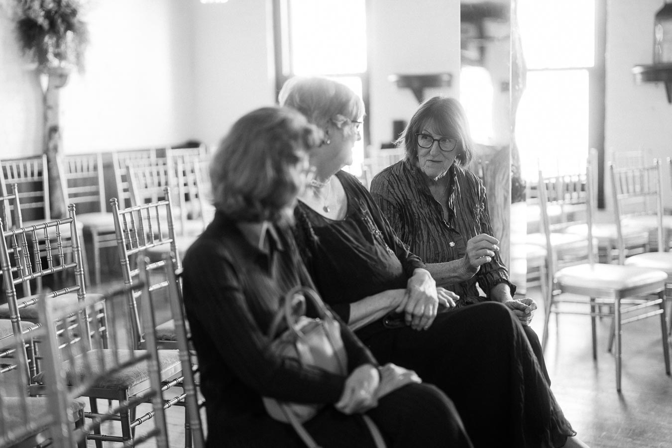 Black and white photograph of three women seated in a softly lit room with rows of empty chairs, engaged in conversation.