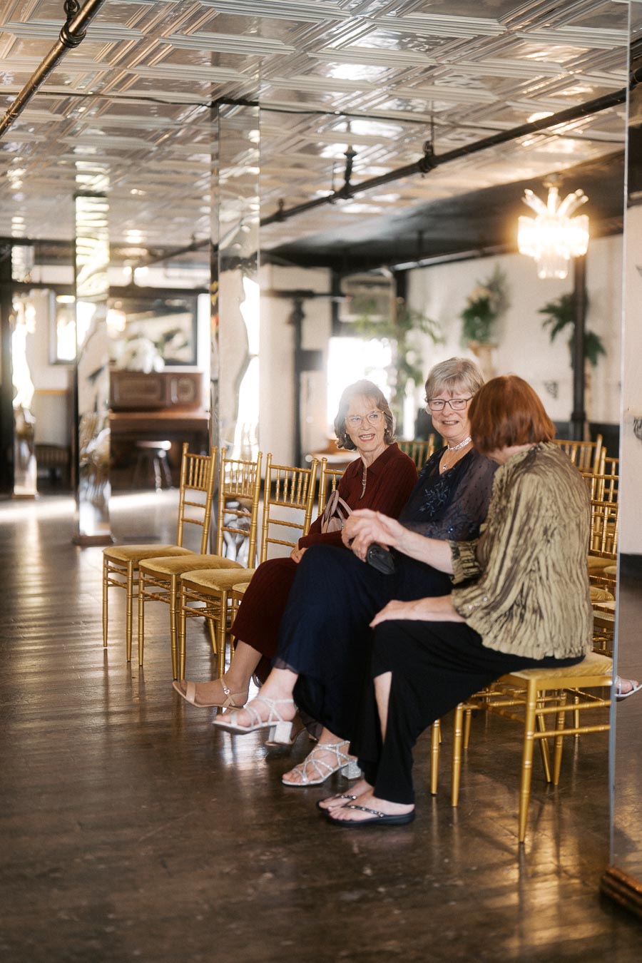 Elderly women sitting on golden chairs in a vintage-style room with mirrored walls and wooden flooring, engaging in conversation and enjoying a social gathering.