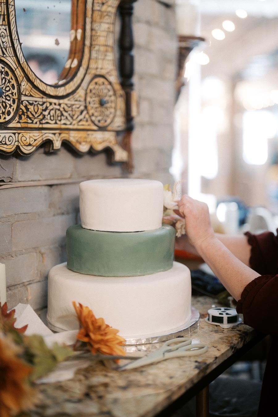 Three-tiered wedding cake being decorated with flowers, set on a marble counter with an ornate mirror in the background.