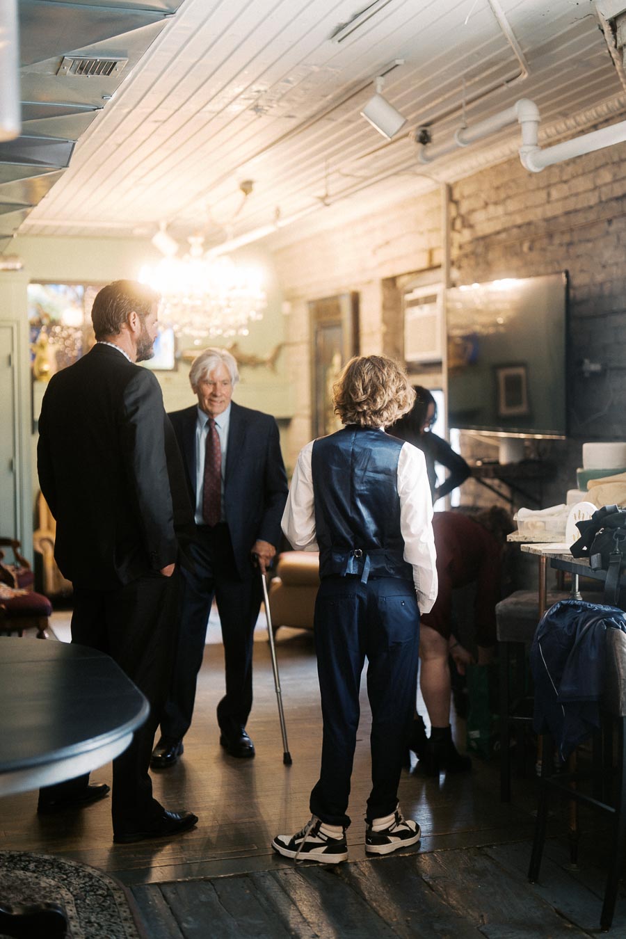 A group of elegantly dressed people, including an elderly man with a cane, gathers in a warmly lit vintage-style room with wooden floors and exposed brick walls, conveying a sense of timeless sophistication and intimate conversation.