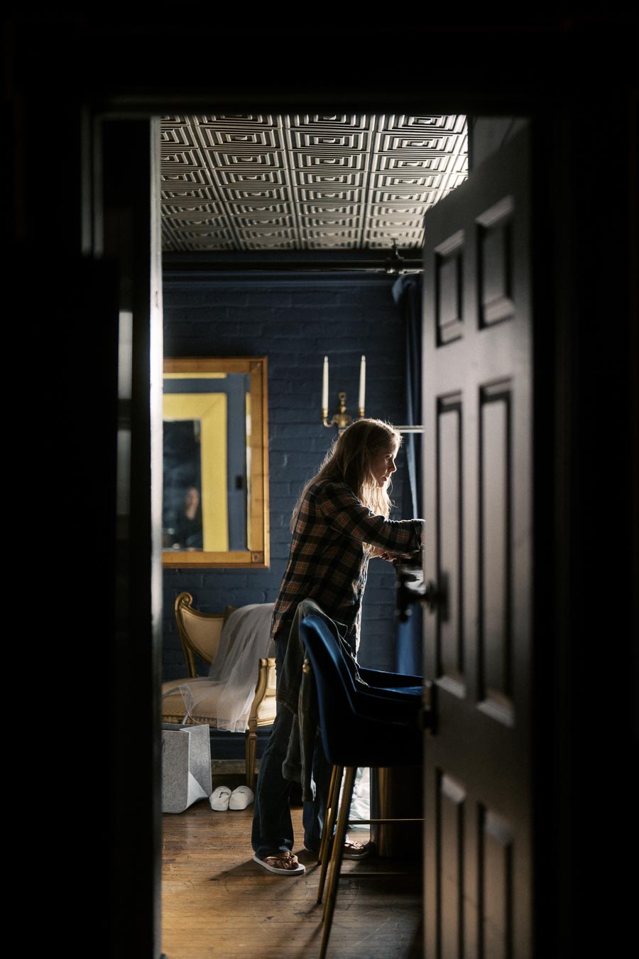 A person standing in a dimly lit room, arranging items on a table with a mirror reflecting the scene. The setting features modern decor with a patterned ceiling and a blue wall.