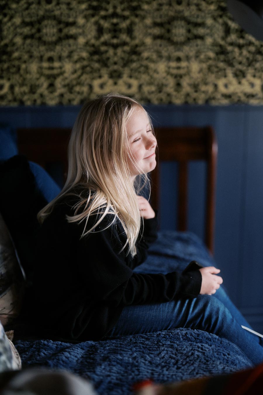 A young girl with long blond hair sitting on a bed, smiling with eyes closed, in a cozy room with dark blue walls and patterned wallpaper.