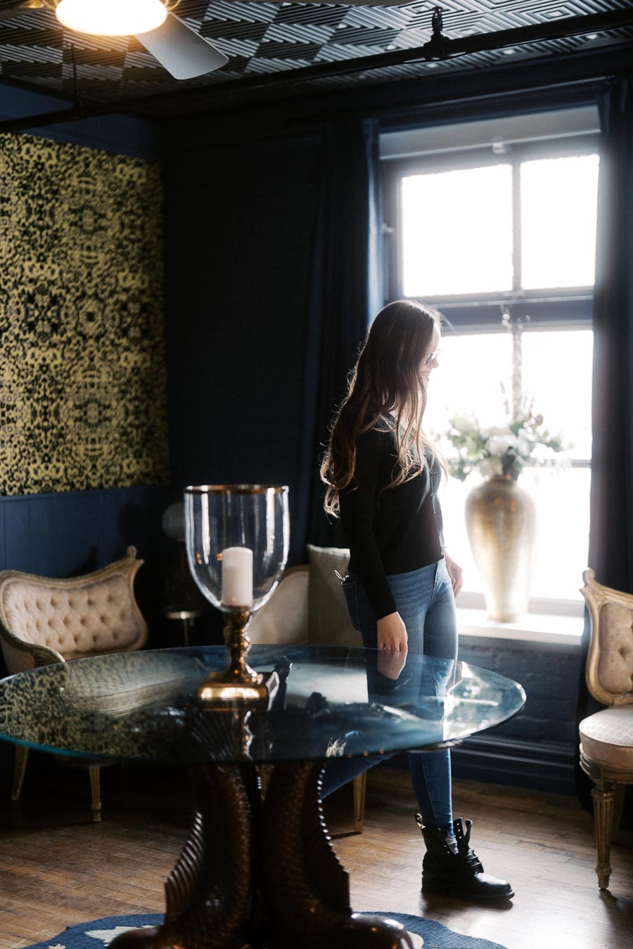 Elegant interior with a person standing by a sunlit window, decorated with ornate furniture, a glass table, and a stylish vase.