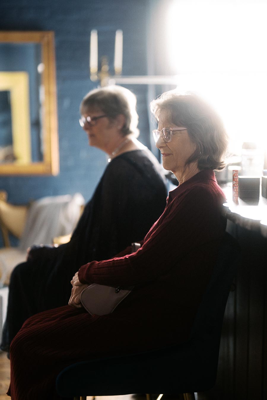 Elderly women sitting in a dimly lit room, with soft natural light illuminating them, wearing glasses and casual attire, conveying a peaceful and reflective atmosphere.