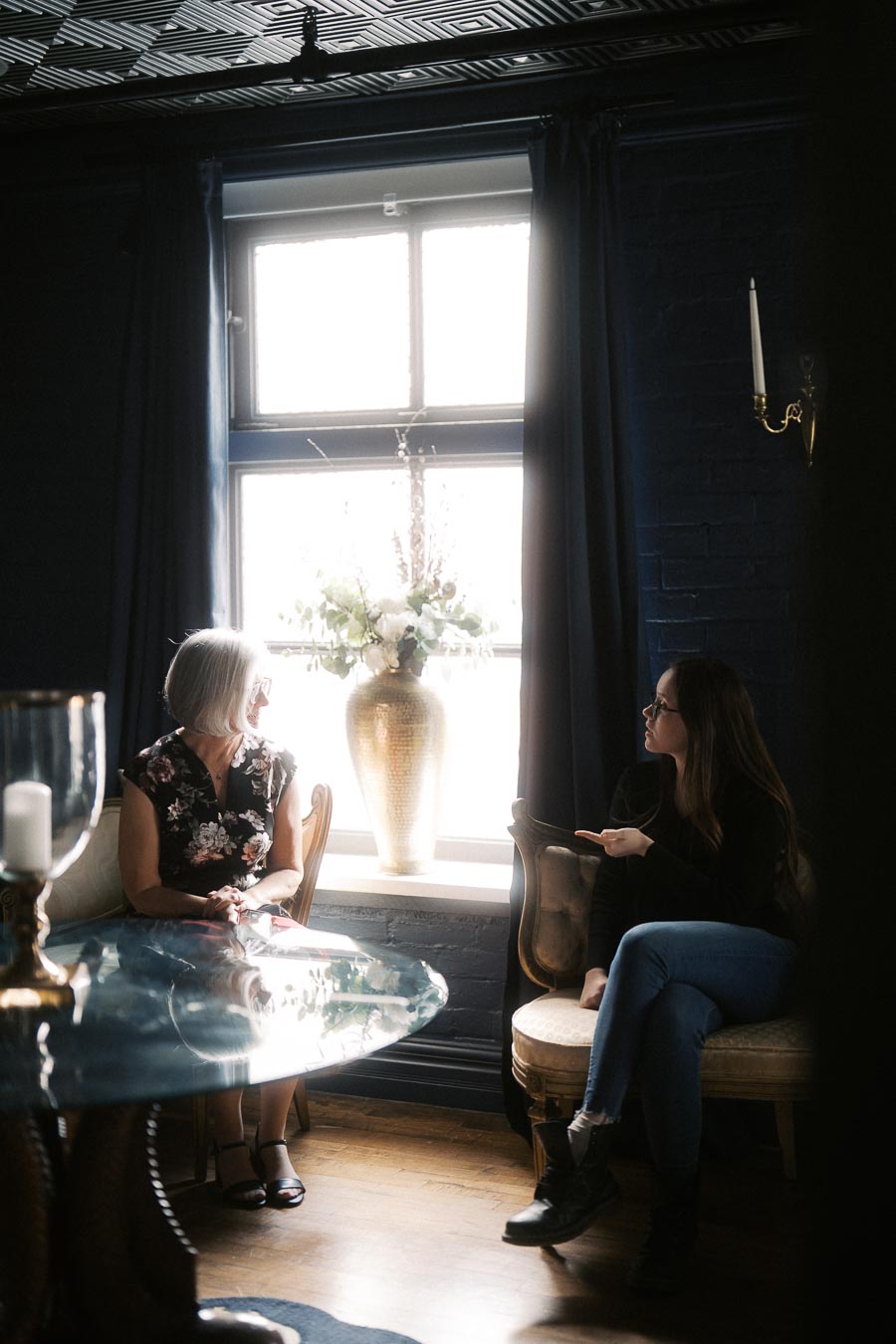 Two women sitting in a softly lit room, engaging in conversation. A large window with sheer curtains illuminates the space, highlighting a decorative vase with flowers on the windowsill. The environment is cozy and elegant, featuring a dark blue wall and a glass-top table.