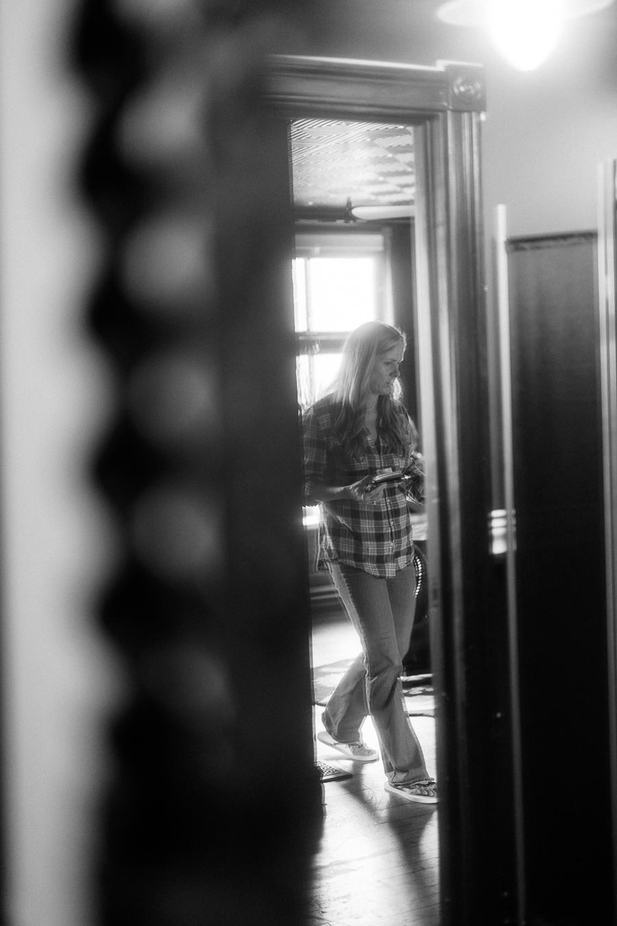 Black and white image of a woman in a plaid shirt holding an object, captured through a doorway, with soft lighting creating a vintage ambiance.