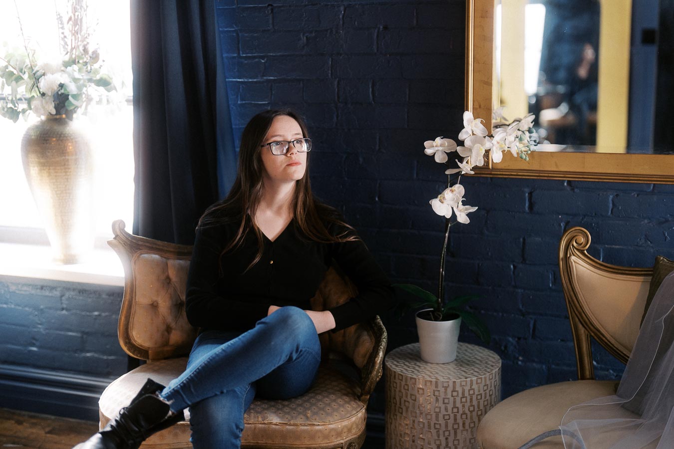 A woman sits thoughtfully on a vintage chair in a stylishly decorated room with dark blue brick walls. She is wearing glasses, a black top, and jeans. Beside her is a small table with a potted white orchid, and a large mirror hangs on the wall. Soft natural light enters from a window adorned with flowers.