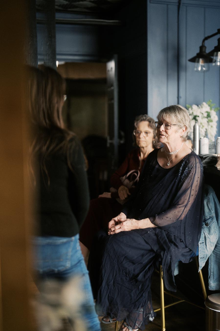 An elderly woman in a navy dress sitting and engaging in conversation indoors, with another elderly woman in the background and a person standing nearby. The room features dark blue walls and soft lighting.
