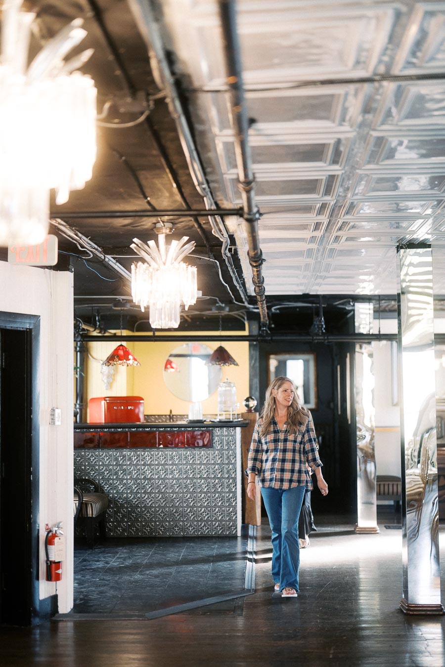 A woman in casual attire walks through a stylish, vintage-themed interior with ornate lighting and decorative ceilings, featuring a retro-style bar and mirror accents.