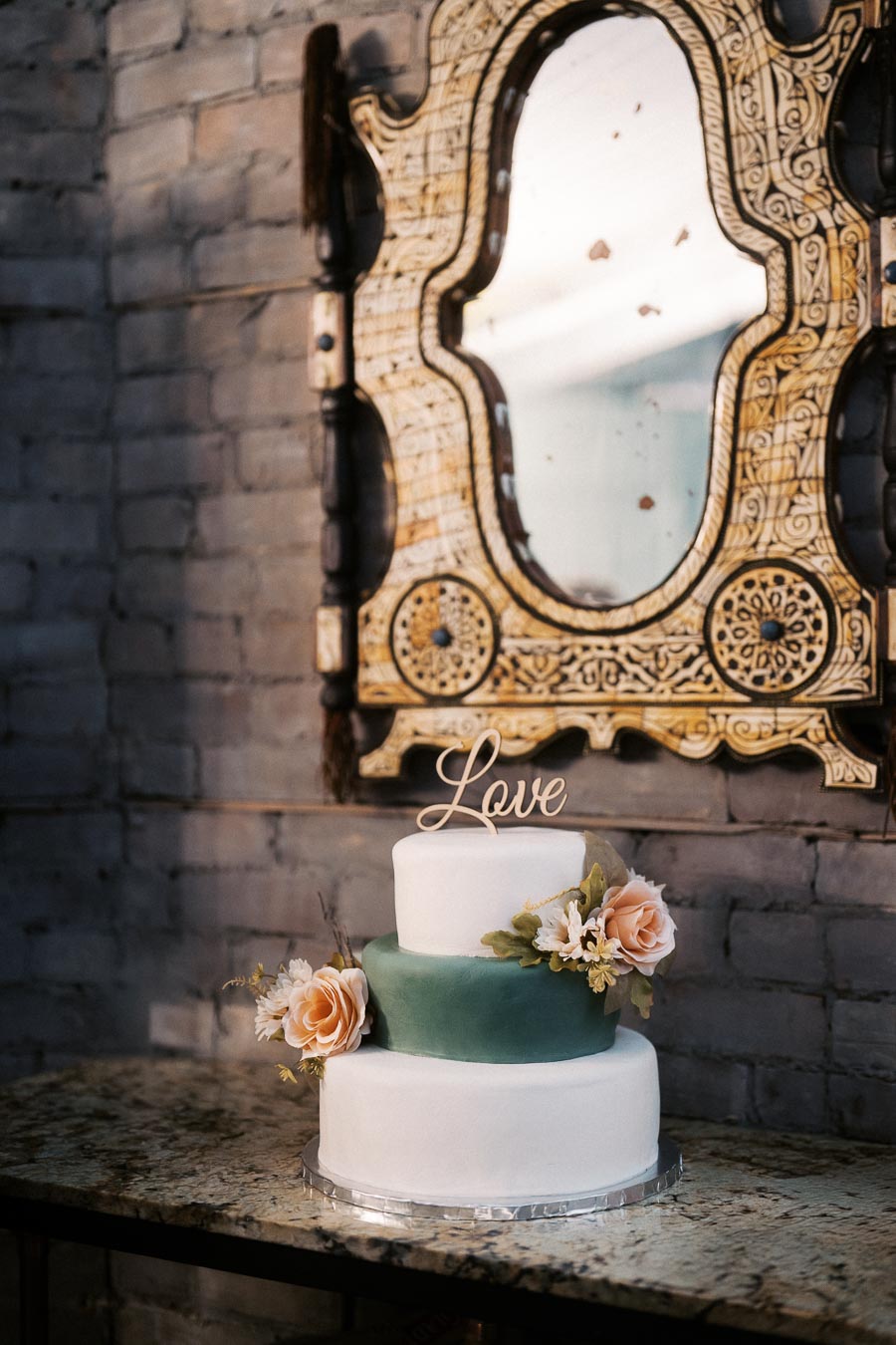 Elegant two-tier wedding cake adorned with ivory and pink floral decorations, set in a rustic setting with a vintage mirror, topped with a wooden Love sign, capturing a romantic and timeless atmosphere.